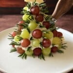 A festive appetizer shaped like a Christmas tree made of green and red grapes, sprigs of rosemary, and cheese, topped with a star-shaped cheese piece, on a white plate with a cheese knife.