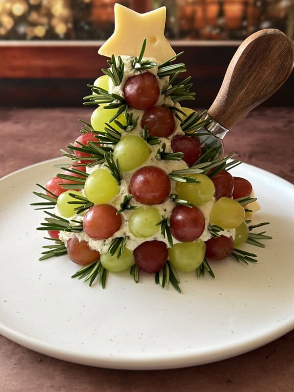 A festive appetizer shaped like a Christmas tree made of green and red grapes, sprigs of rosemary, and cheese, topped with a star-shaped cheese piece, on a white plate with a cheese knife.