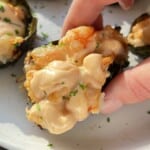 A hand holds a piece of baked sushi topped with creamy sauce and herbs, with more sushi pieces on a white plate in the background.
