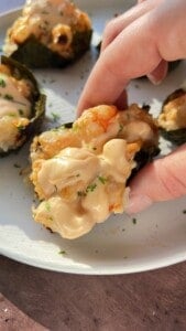 A hand holds a piece of baked sushi topped with creamy sauce and herbs, with more sushi pieces on a white plate in the background.