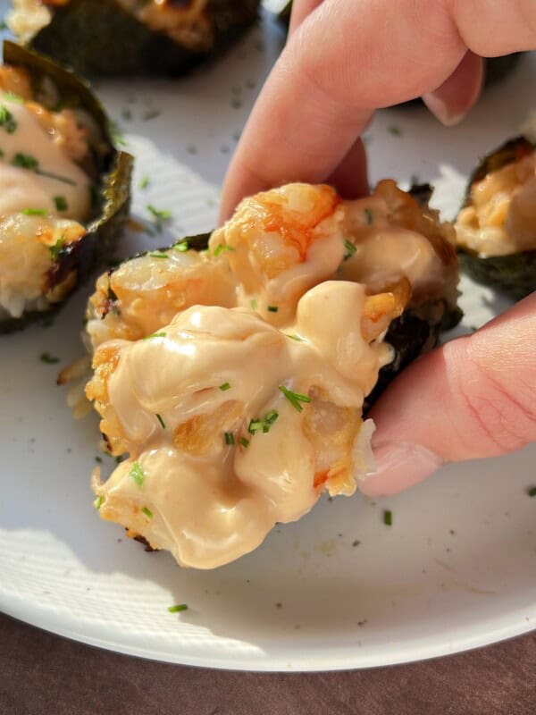 A hand holds a piece of baked sushi topped with creamy sauce and herbs, with more sushi pieces on a white plate in the background.