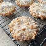 Close-up of pine nut cookies dusted with powdered sugar, cooling on a black wire rack over parchment paper. Several cookies are visible, with pine nuts covering their surfaces.