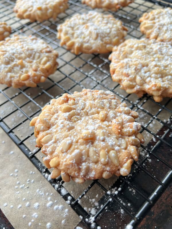 Close-up of pine nut cookies dusted with powdered sugar, cooling on a black wire rack over parchment paper. Several cookies are visible, with pine nuts covering their surfaces.