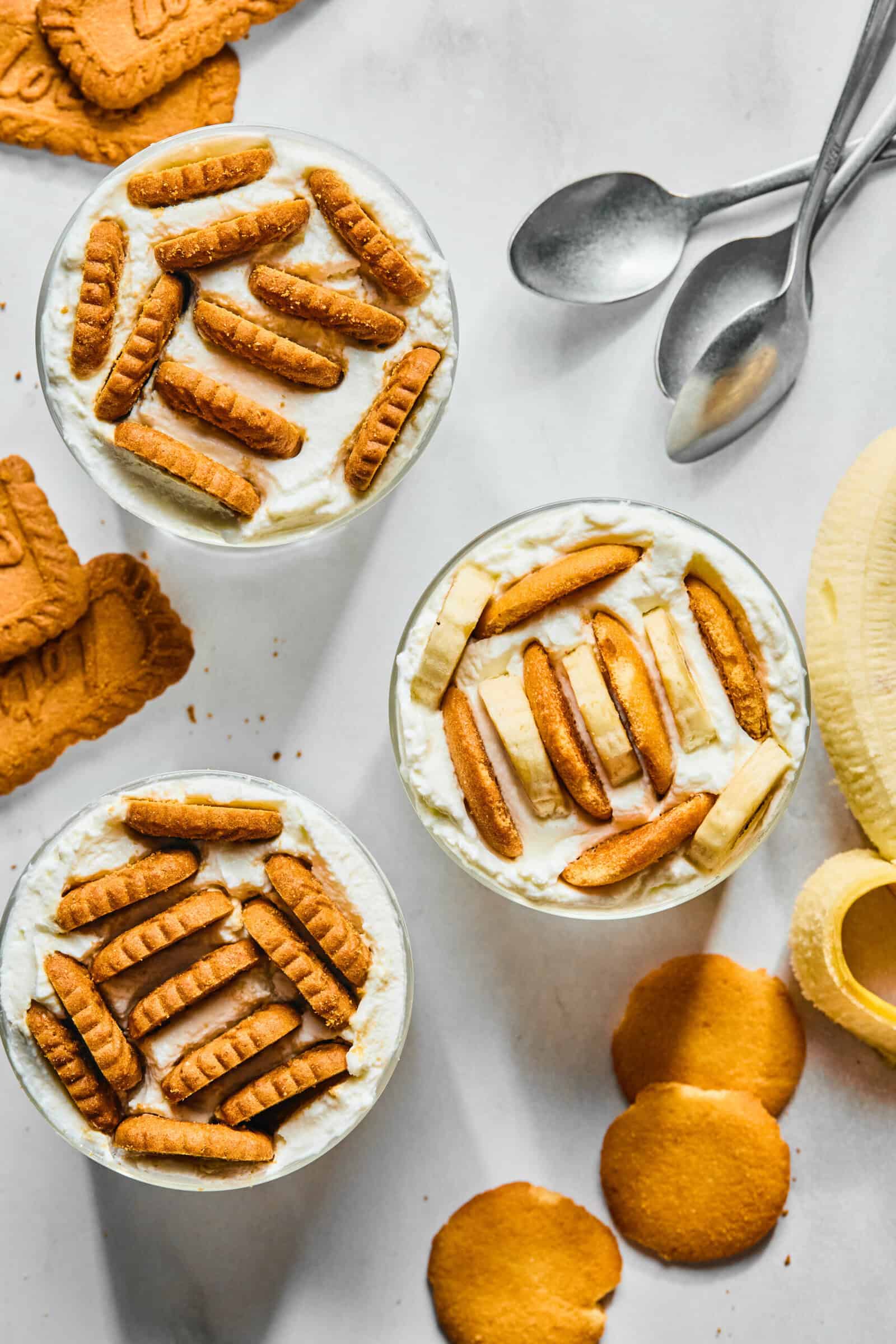 Three dessert cups topped with cookies and banana slices are arranged on a white surface, inspired by the Viral Japanese Yogurt Cheesecake trend, surrounded by extra cookies, banana slices, and three metal spoons. - 4