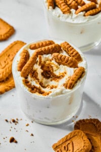 A glass filled with creamy white Viral Japanese Yogurt Cheesecake dessert, topped and swirled with crumbled and whole Biscoff cookies. Cookie pieces and crumbs are scattered on the white surface around the glass.