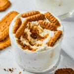A glass filled with creamy dessert inspired by Viral Japanese Yogurt Cheesecake, topped with crumbled and whole caramelized Biscoff cookies. Additional cookies and crumbs are scattered on the white surface around the glass.