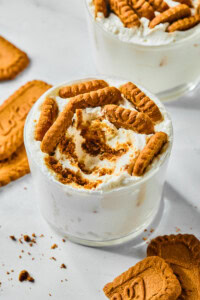 A glass filled with creamy dessert inspired by Viral Japanese Yogurt Cheesecake, topped with crumbled and whole caramelized Biscoff cookies. Additional cookies and crumbs are scattered on the white surface around the glass.