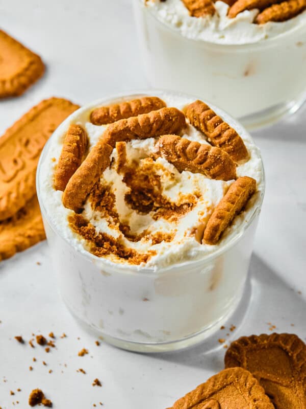 A glass filled with creamy dessert inspired by Viral Japanese Yogurt Cheesecake, topped with crumbled and whole caramelized Biscoff cookies. Additional cookies and crumbs are scattered on the white surface around the glass.