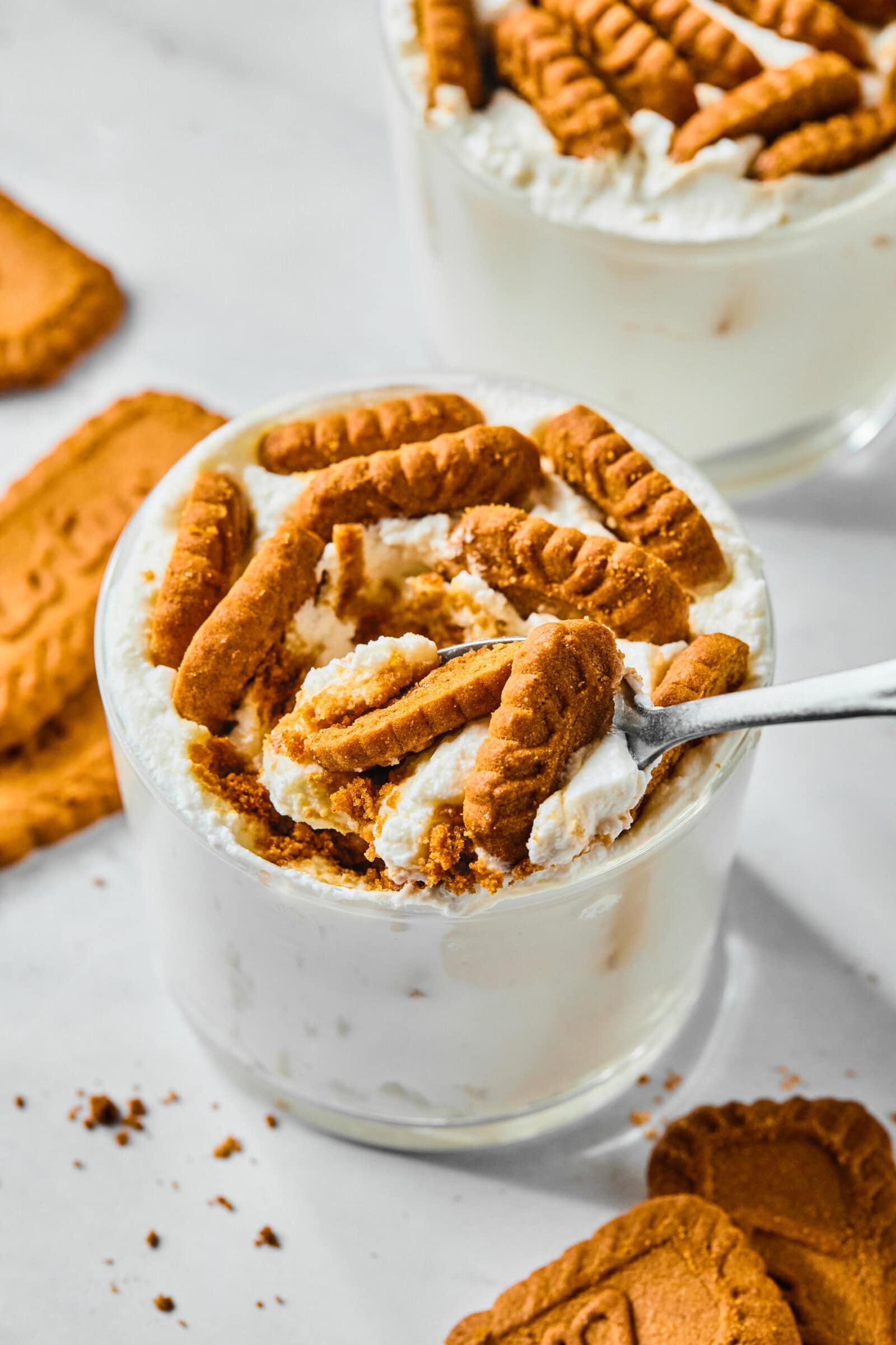 A glass of creamy dessert inspired by the viral Japanese Yogurt Cheesecake, topped with crushed and whole Biscoff cookies. A spoon scoops some out, while cookie crumbs are scattered on the white surface nearby. - 3
