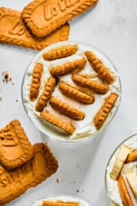 A glass filled with creamy dessert inspired by Viral Japanese Yogurt Cheesecake, topped with Lotus Biscoff cookies, with additional cookies scattered around on a white surface.