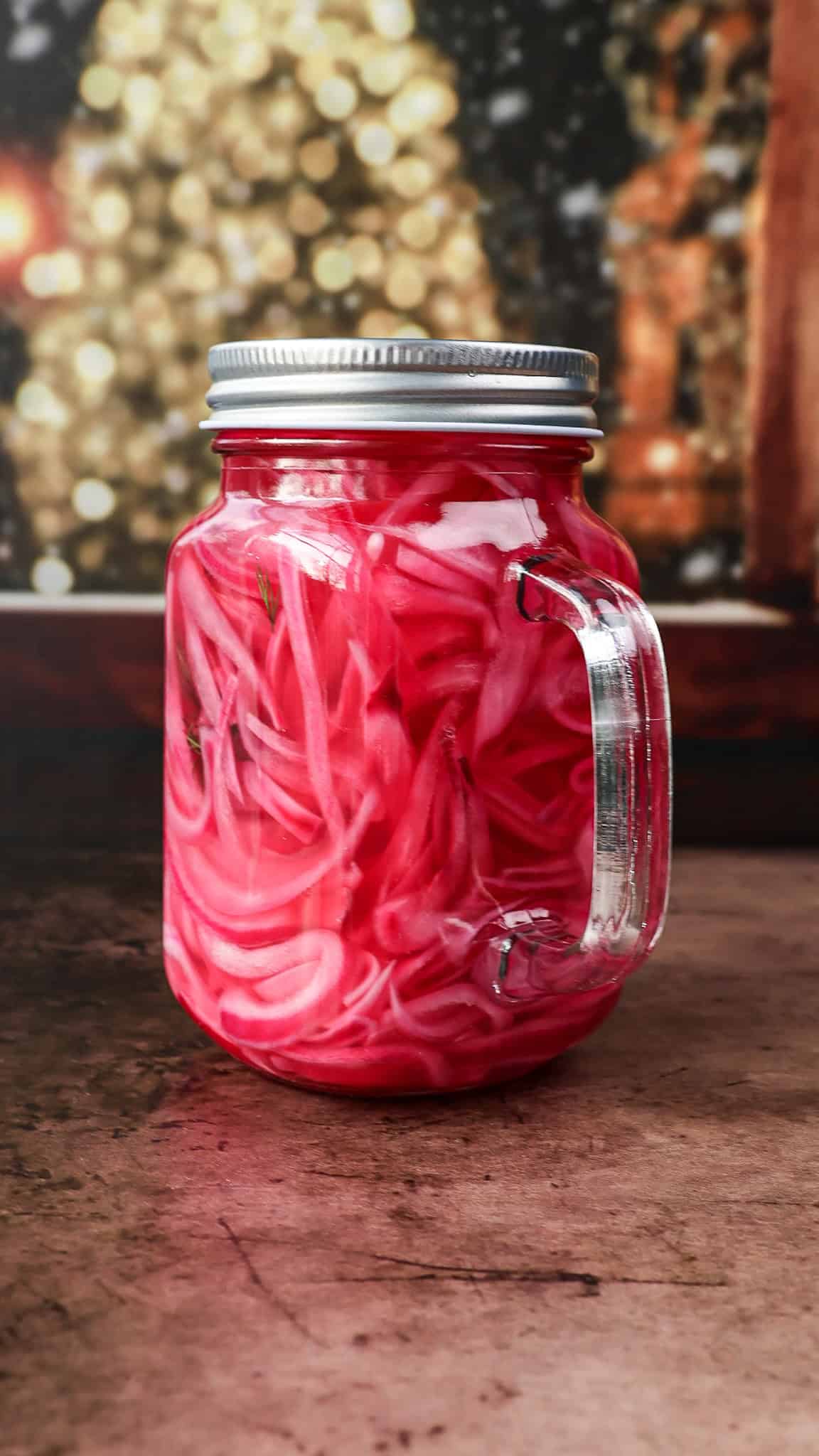 A glass jar with a handle, filled with bright pink pickled red onions, sits on a rustic surface with a blurred, festive background of warm lights. - 4