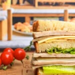 A stacked sandwich with layers of lettuce, cheese, and deli meat on thick bread slices sits on a rustic paper with tomatoes on the vine nearby and a bakery display blurred in the background.