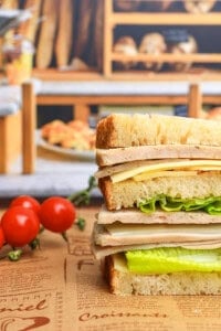 A stacked sandwich with layers of lettuce, cheese, and deli meat on thick bread slices sits on a rustic paper with tomatoes on the vine nearby and a bakery display blurred in the background.