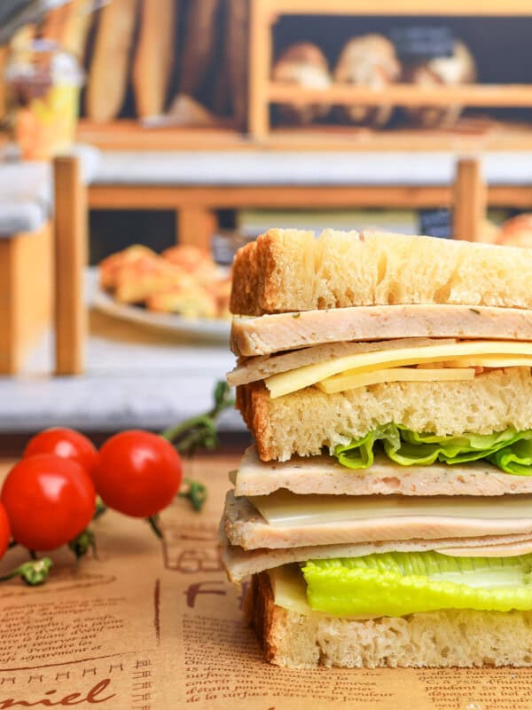 A stacked sandwich with layers of lettuce, cheese, and deli meat on thick bread slices sits on a rustic paper with tomatoes on the vine nearby and a bakery display blurred in the background.