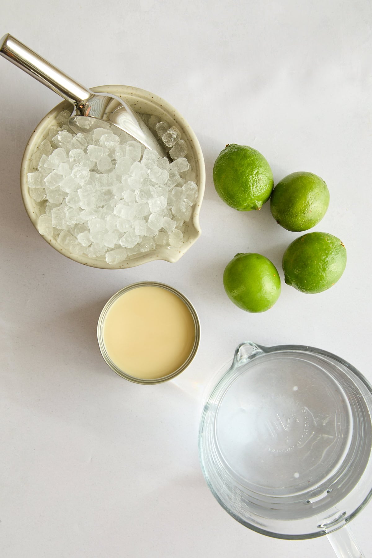 A top-down view of a bowl of crushed ice with a scoop, three whole limes, an open can of sweetened condensed milk, and a glass pitcher of water on a white surface. - 4