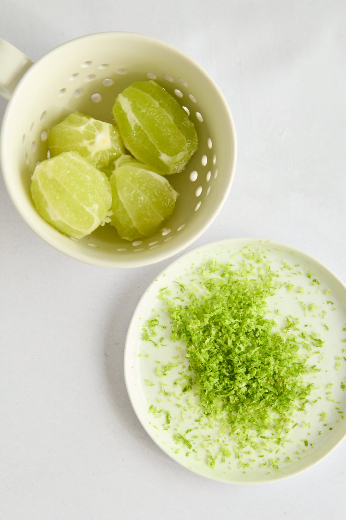A mug with peeled limes sits above a small plate holding a pile of fresh lime zest, all on a light surface.