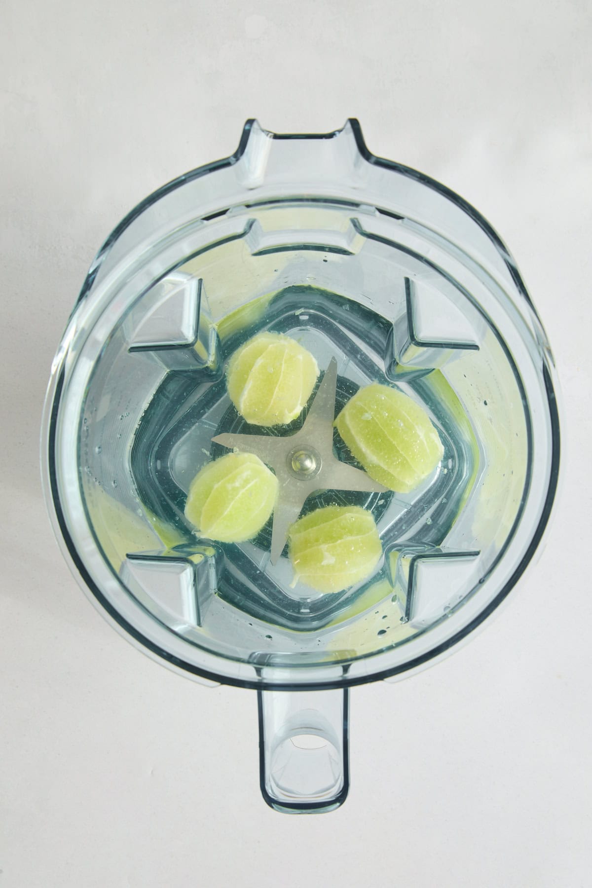 Top view of a blender with four peeled lime halves and water inside, placed on a light-colored surface.