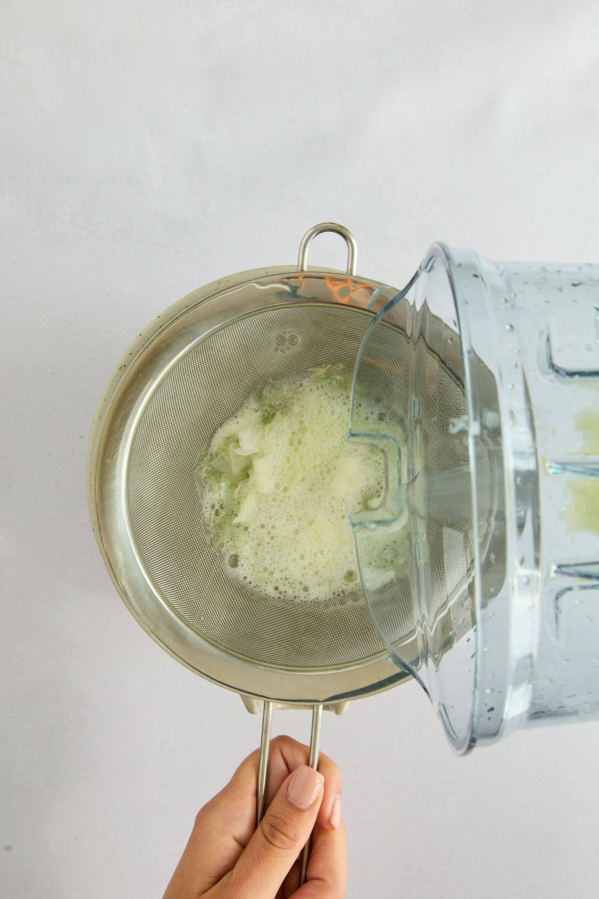 A hand holds a metal strainer over a bowl while pouring a foamy liquid from a blender through the strainer, separating pulp from juice. - 5