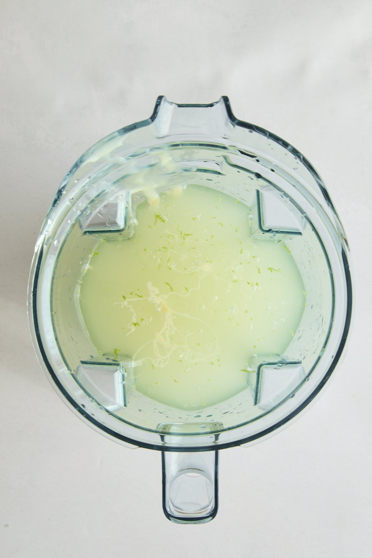 Top-down view of a blender containing Brazilian Lemonade - a pale green liquid with small flecks of lime zest, set against a light background.