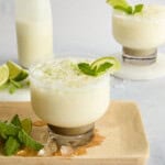 A creamy white drink garnished with lime and mint is served in a glass on a wooden board, with lime wedges, fresh mint, ice cubes, and a milk bottle in the background.
