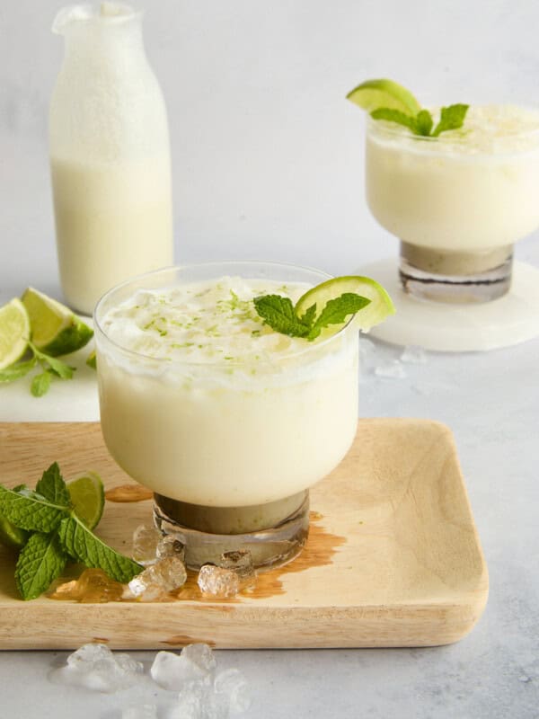 A creamy white drink garnished with lime and mint is served in a glass on a wooden board, with lime wedges, fresh mint, ice cubes, and a milk bottle in the background.
