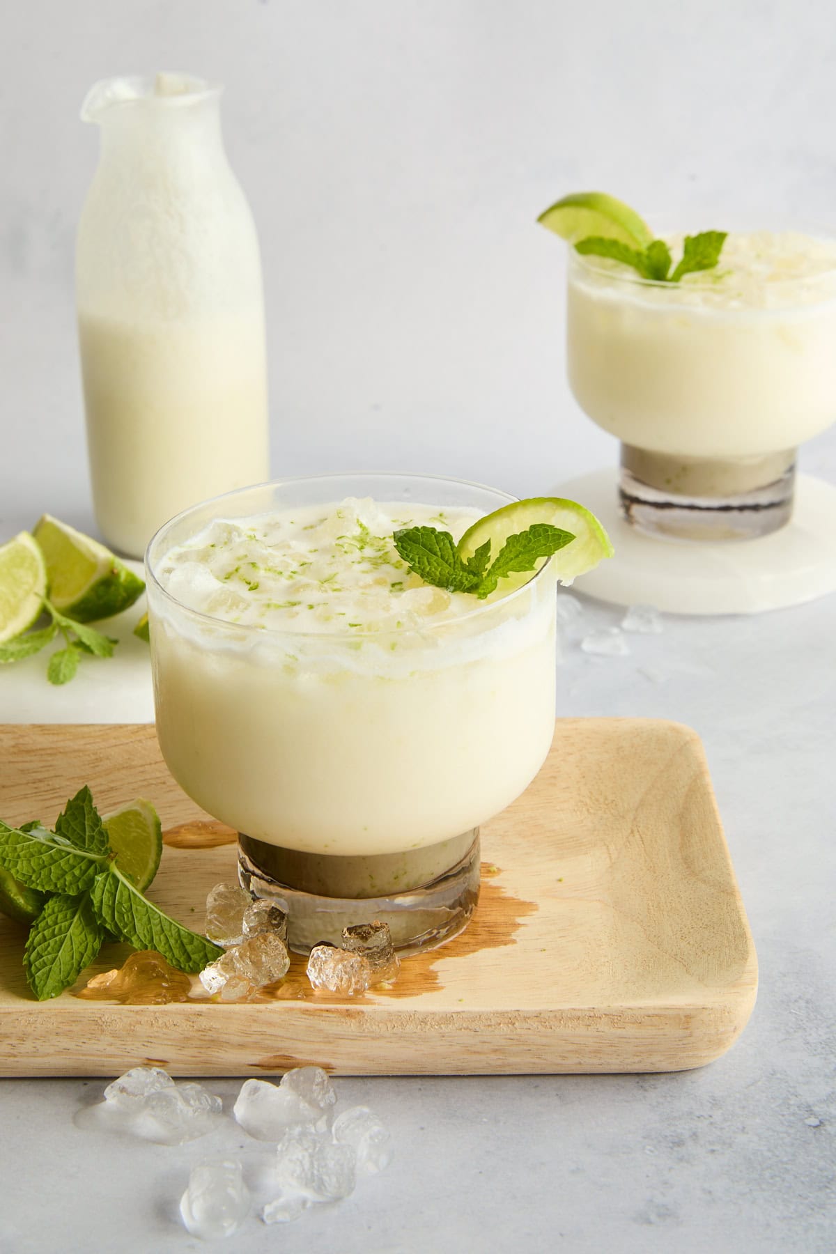 A creamy white drink garnished with lime and mint sits on a wooden tray with ice cubes, lime wedges, and mint leaves. In the background, there’s a glass bottle and another drink on a white surface. - 3