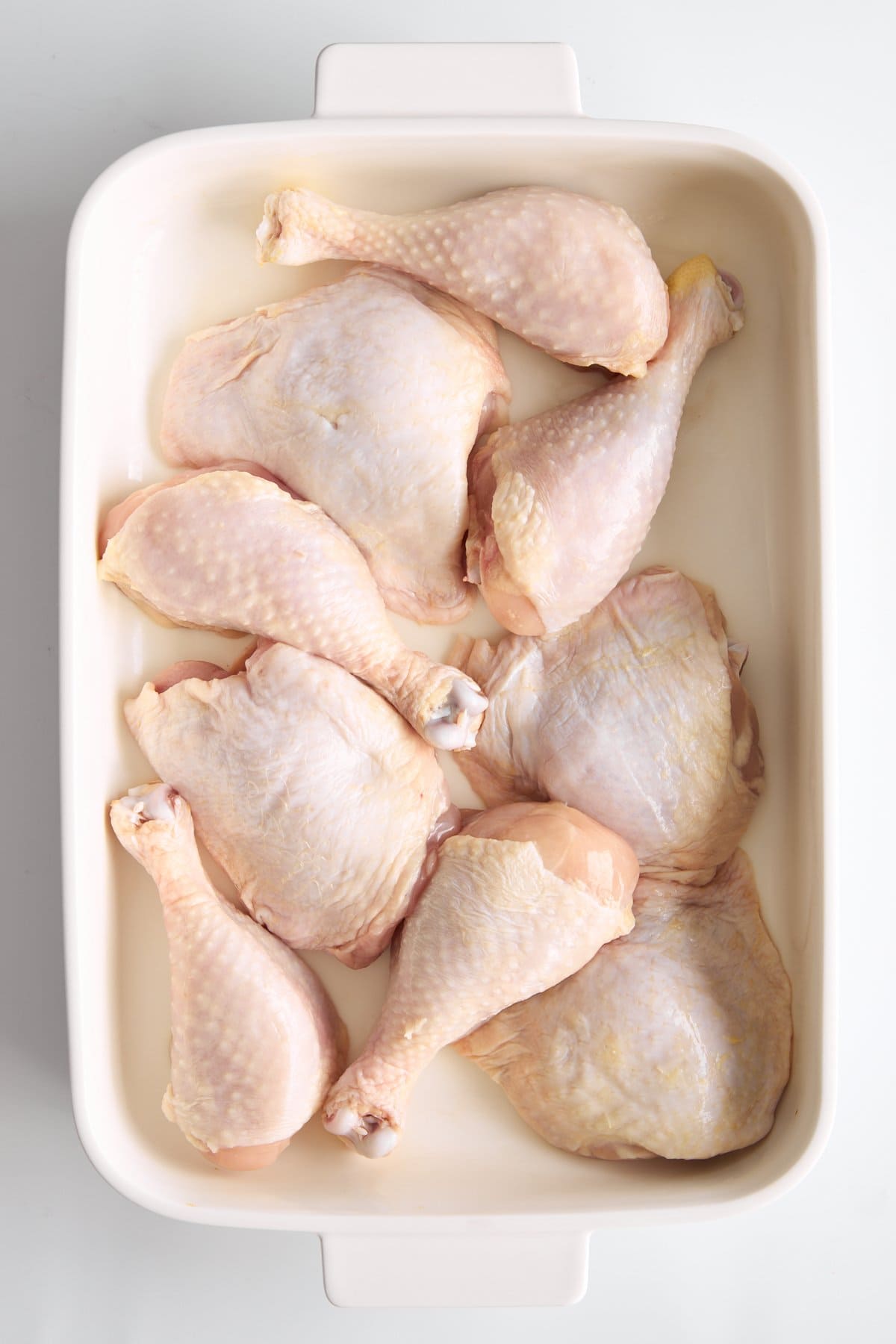 Raw chicken drumsticks and thighs arranged in a single layer inside a white rectangular baking dish on a white surface. - 3