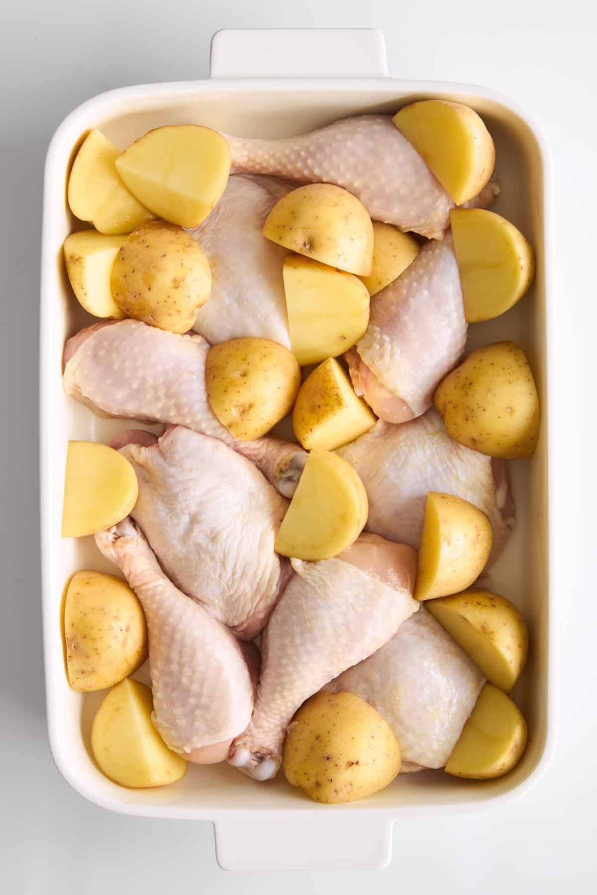 Raw chicken drumsticks and halved yellow potatoes arranged in a white rectangular baking dish, ready to be cooked.