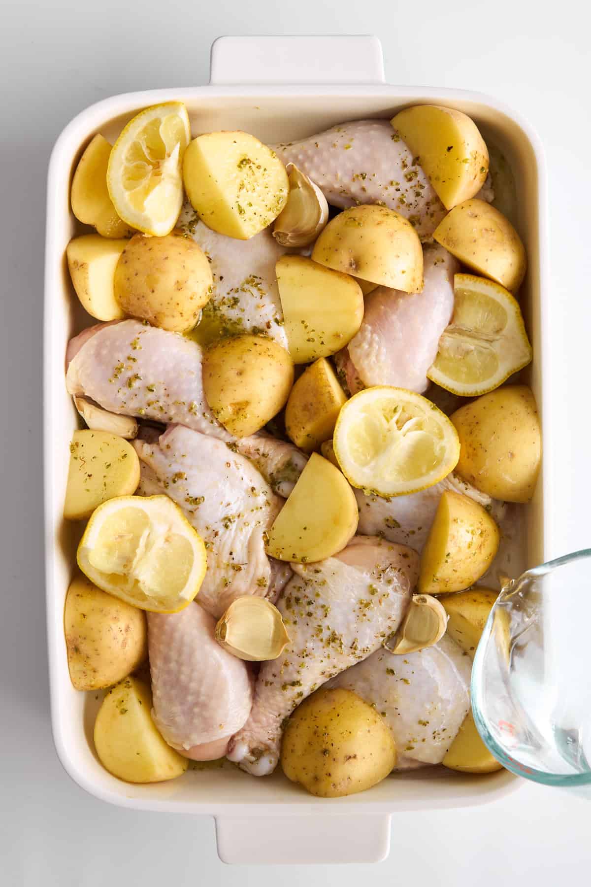 Raw chicken drumsticks, halved yellow potatoes, lemon wedges, and garlic cloves are arranged in a white baking dish, sprinkled with herbs, ready to be cooked.