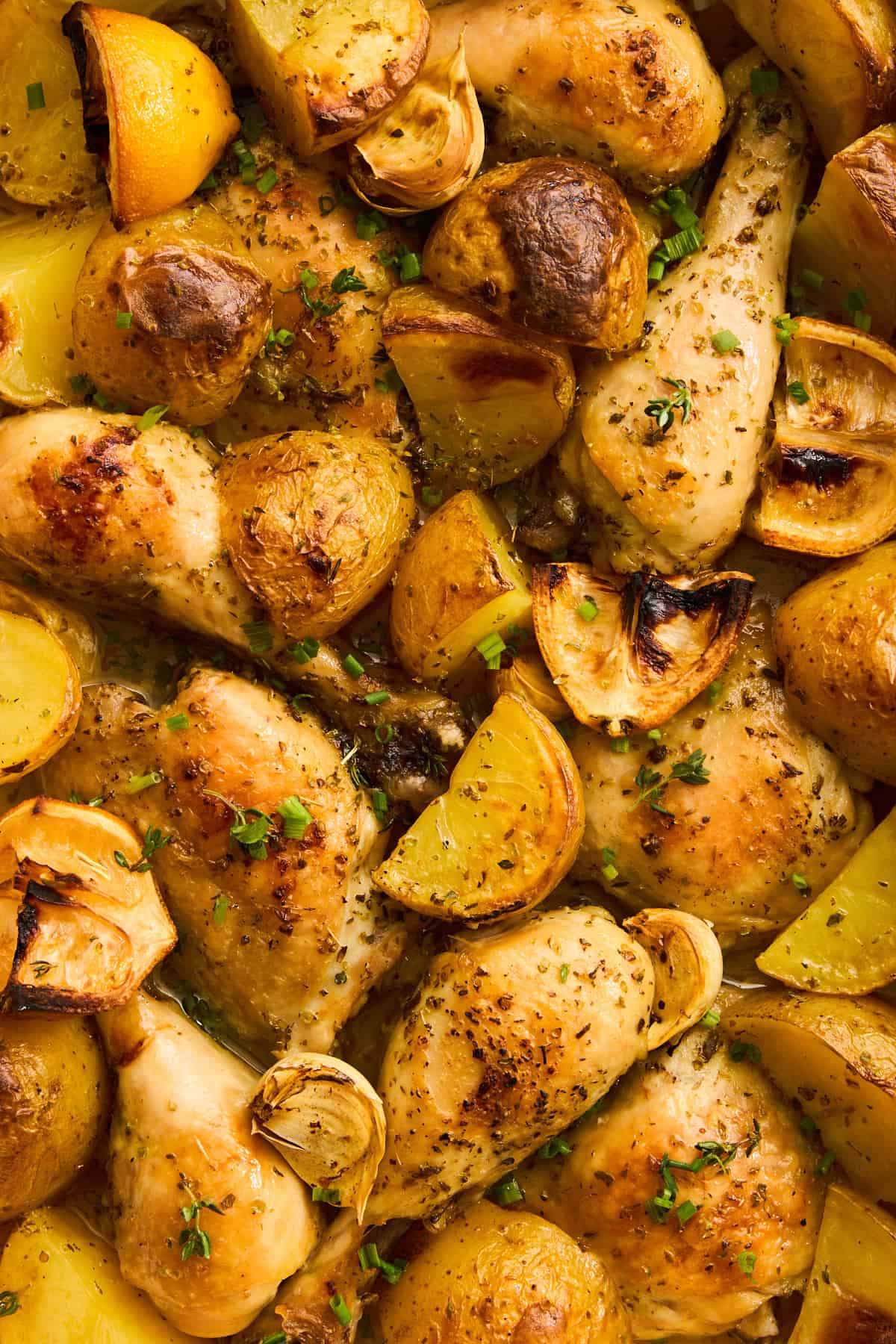 Roasted chicken drumsticks with golden potatoes, lemon wedges, and garlic cloves, all seasoned with herbs and garnished with chopped chives. The dish appears freshly baked and appetizing.