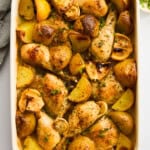 A white baking dish filled with roasted chicken drumsticks, potato wedges, and garlic cloves, garnished with herbs. The dish sits on a white surface with a lemon slice, chopped herbs, and a bowl of pepper nearby.