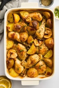 A white baking dish filled with roasted chicken drumsticks, potato wedges, and garlic cloves, garnished with herbs. The dish sits on a white surface with a lemon slice, chopped herbs, and a bowl of pepper nearby.