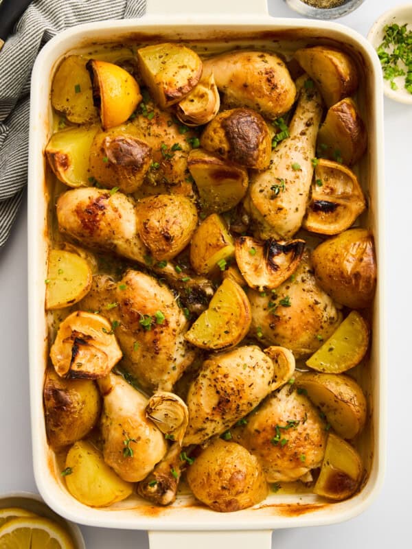 A white baking dish filled with roasted chicken drumsticks, potato wedges, and garlic cloves, garnished with herbs. The dish sits on a white surface with a lemon slice, chopped herbs, and a bowl of pepper nearby. - 7