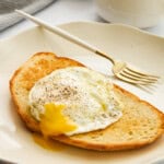 A slice of toasted bread topped with a sunny-side-up egg with a runny yolk, sprinkled with black pepper, on a white plate with a gold fork. A cup of coffee and a striped cloth are in the background.