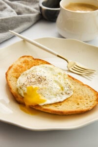 A slice of toasted bread topped with a sunny-side-up egg with a runny yolk, sprinkled with black pepper, on a white plate with a gold fork. A cup of coffee and a striped cloth are in the background.