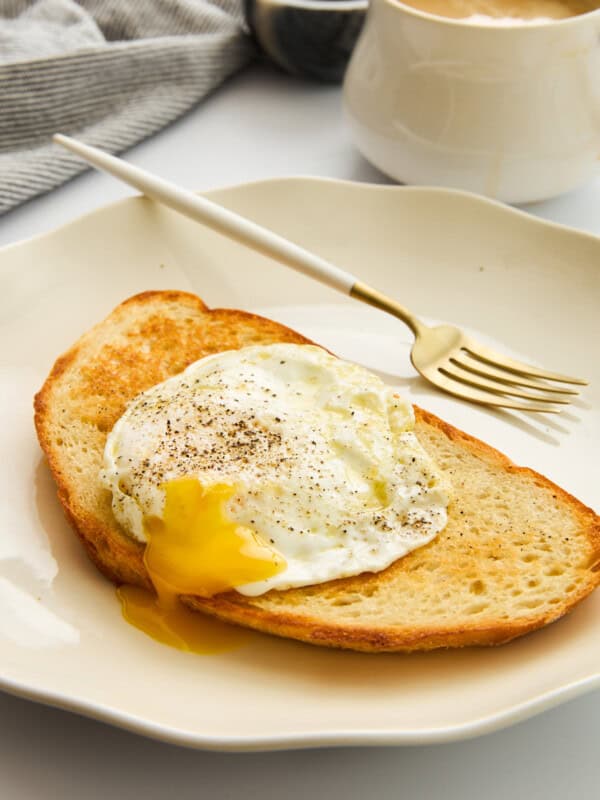 A slice of toasted bread topped with a sunny-side-up egg with a runny yolk, sprinkled with black pepper, on a white plate with a gold fork. A cup of coffee and a striped cloth are in the background.