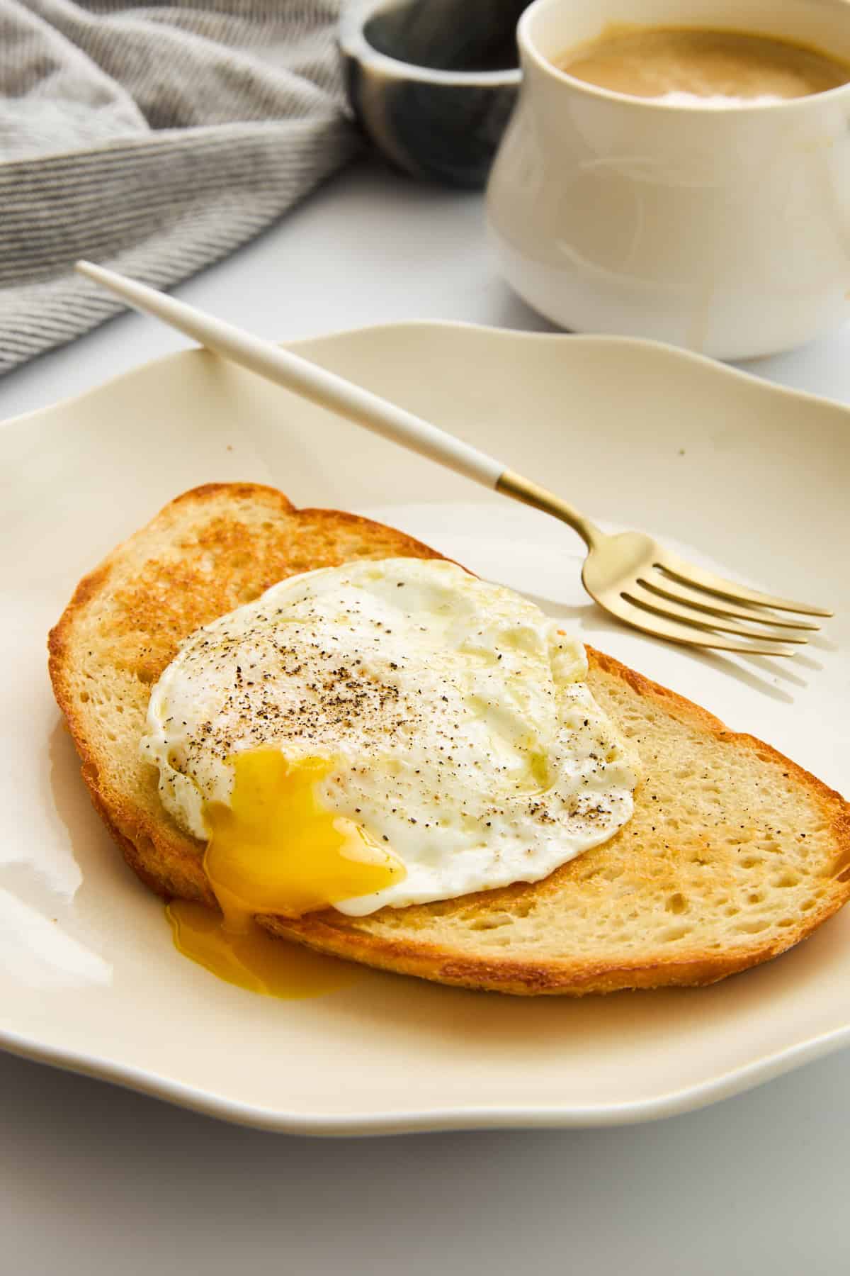 A slice of toasted bread topped with a sunny-side-up egg with a runny yolk, sprinkled with black pepper, on a white plate with a gold fork. A cup of coffee and a striped cloth are in the background. - 4