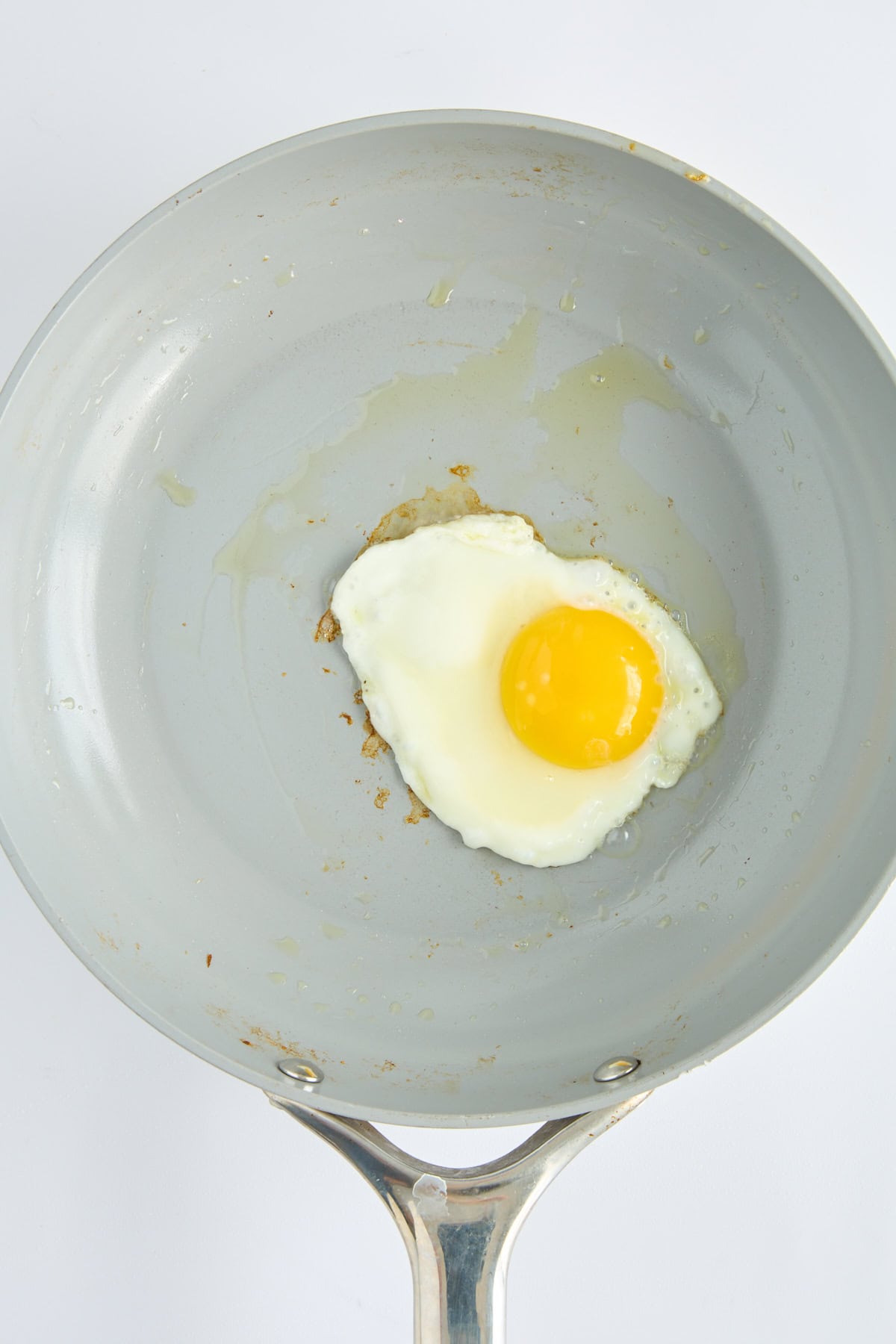 A single sunny-side-up egg frying in a light gray nonstick skillet with a metal handle, seen from above against a white background. - 5