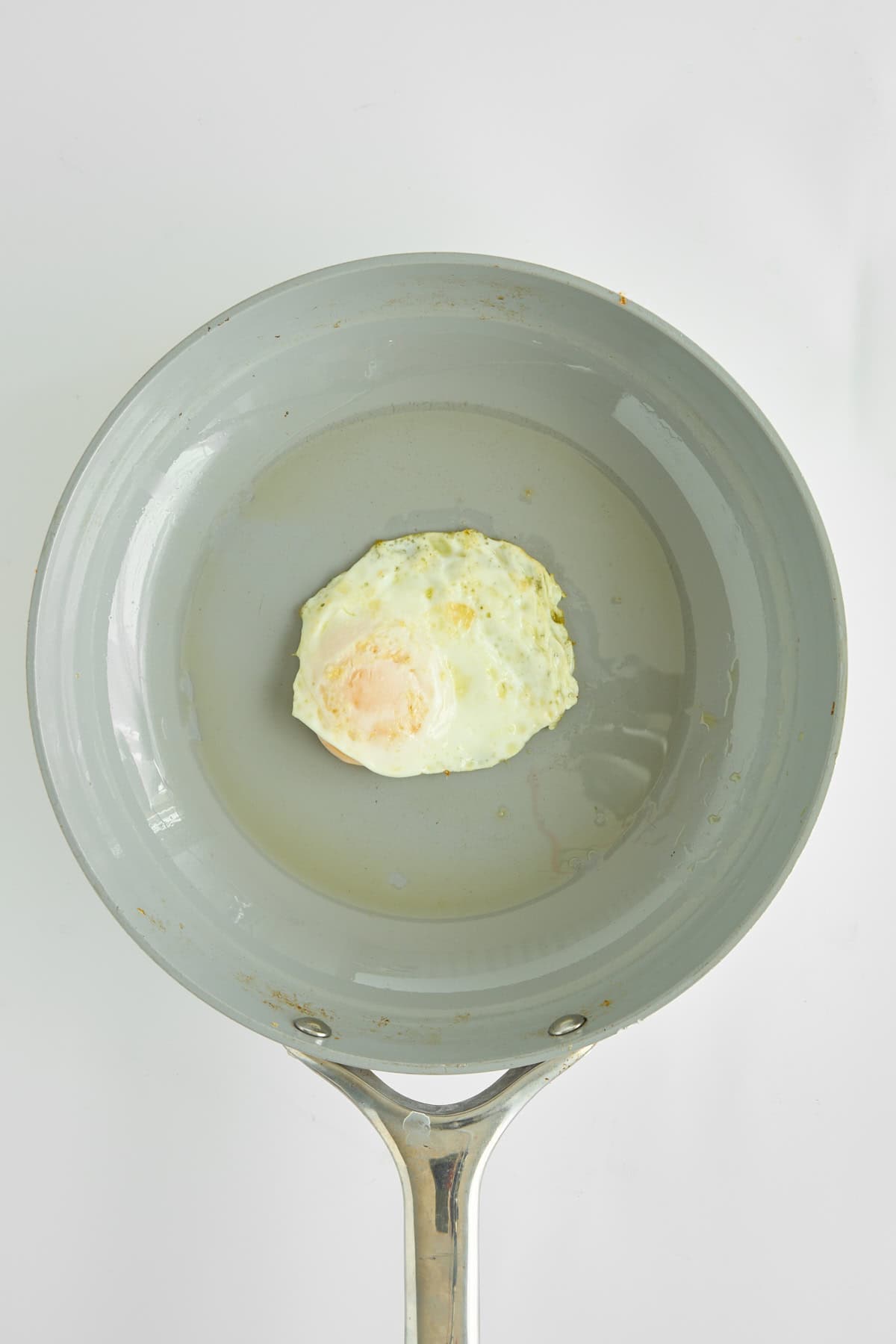 A single fried egg sits in the center of a light gray nonstick frying pan with a metal handle, viewed from above on a white background.