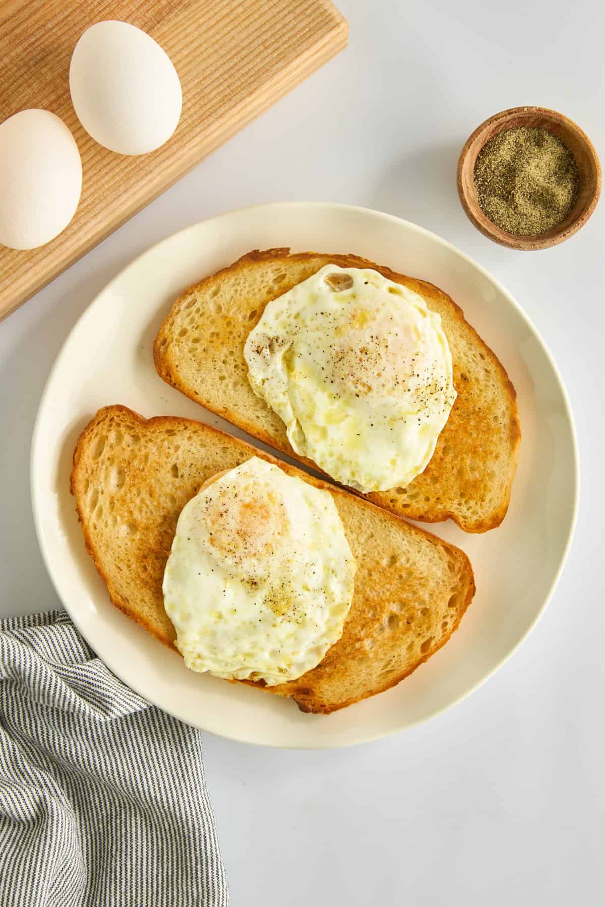 Two slices of toasted bread each topped with a fried egg and sprinkled with pepper on a white plate, next to a small bowl of pepper, two eggs on a board, and a striped kitchen towel. - 3