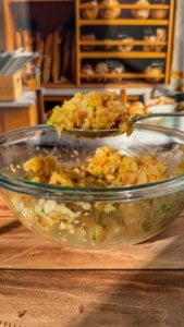 A glass bowl filled with a mixture of rice, vegetables, and herbs sits on a wooden table; a spoon holding a portion of the mixture is lifted above the bowl, with a bakery shelf blurred in the background.