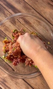 A hand mixing ground meat with chopped vegetables and seasonings in a clear glass bowl on a wooden surface.
