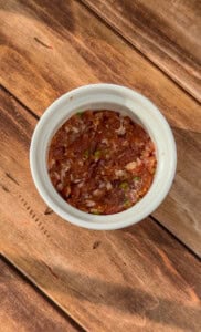 A white bowl filled with a chunky red salsa or dip, with visible green and white vegetable pieces, placed on a rustic wooden surface.