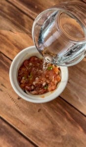 A glass cup pours water into a white bowl filled with a chunky tomato mixture, sitting on a wooden surface.