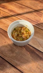 A small white ramekin filled with finely chopped sautéed onions and green peppers sits on a wooden surface in warm sunlight.