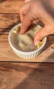 A hand pressing a dumpling wrapper into a white ramekin on a wooden surface, shaping the dough to fit the dish.