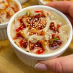 A hand holds a small white ramekin filled with noodles topped with chili oil, sesame seeds, and chopped green onions; three similar ramekins are in the background on a wooden board.