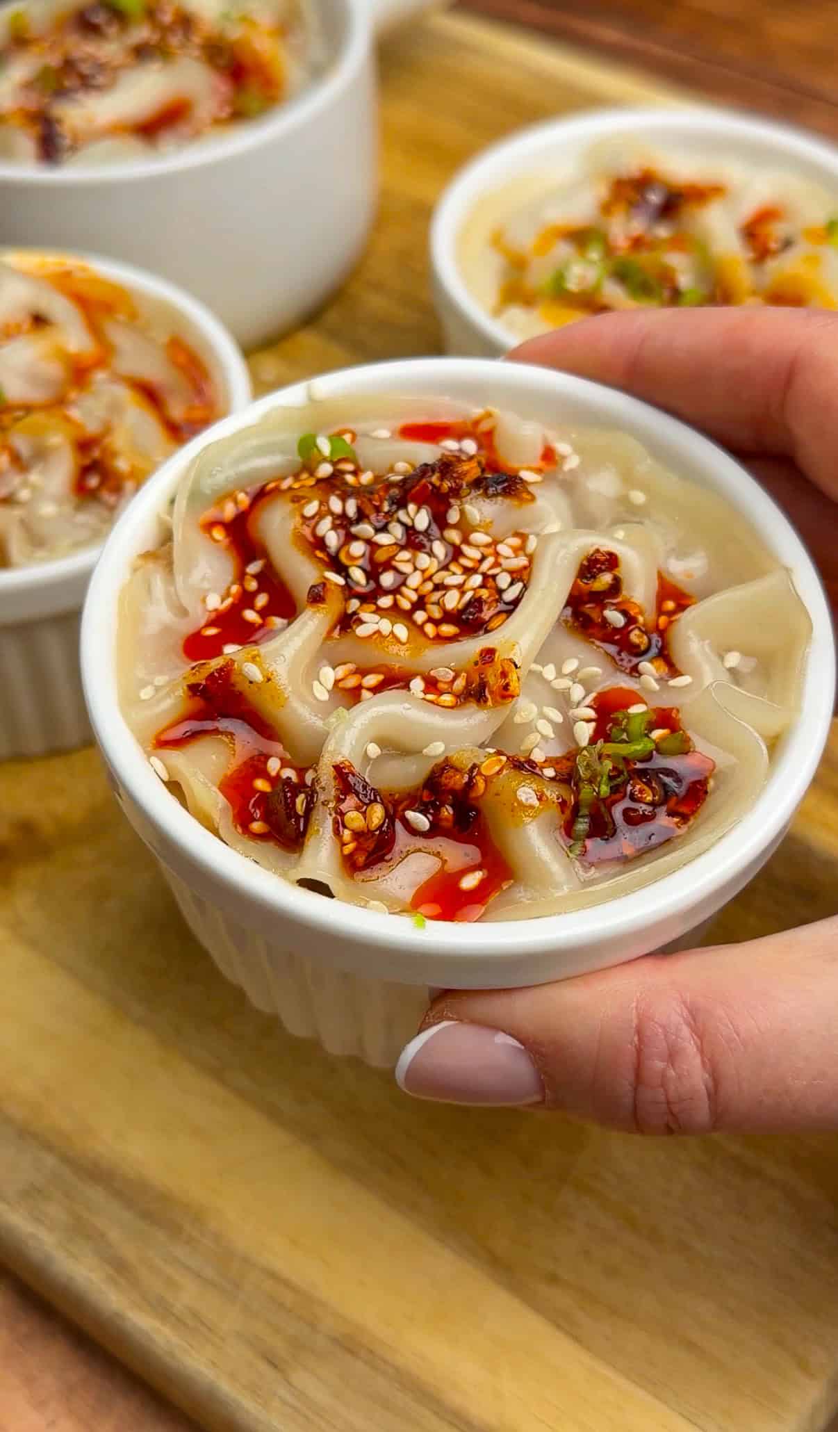 A hand holds a small white ramekin filled with noodles topped with chili oil, sesame seeds, and chopped green onions; three similar ramekins are in the background on a wooden board. - 4