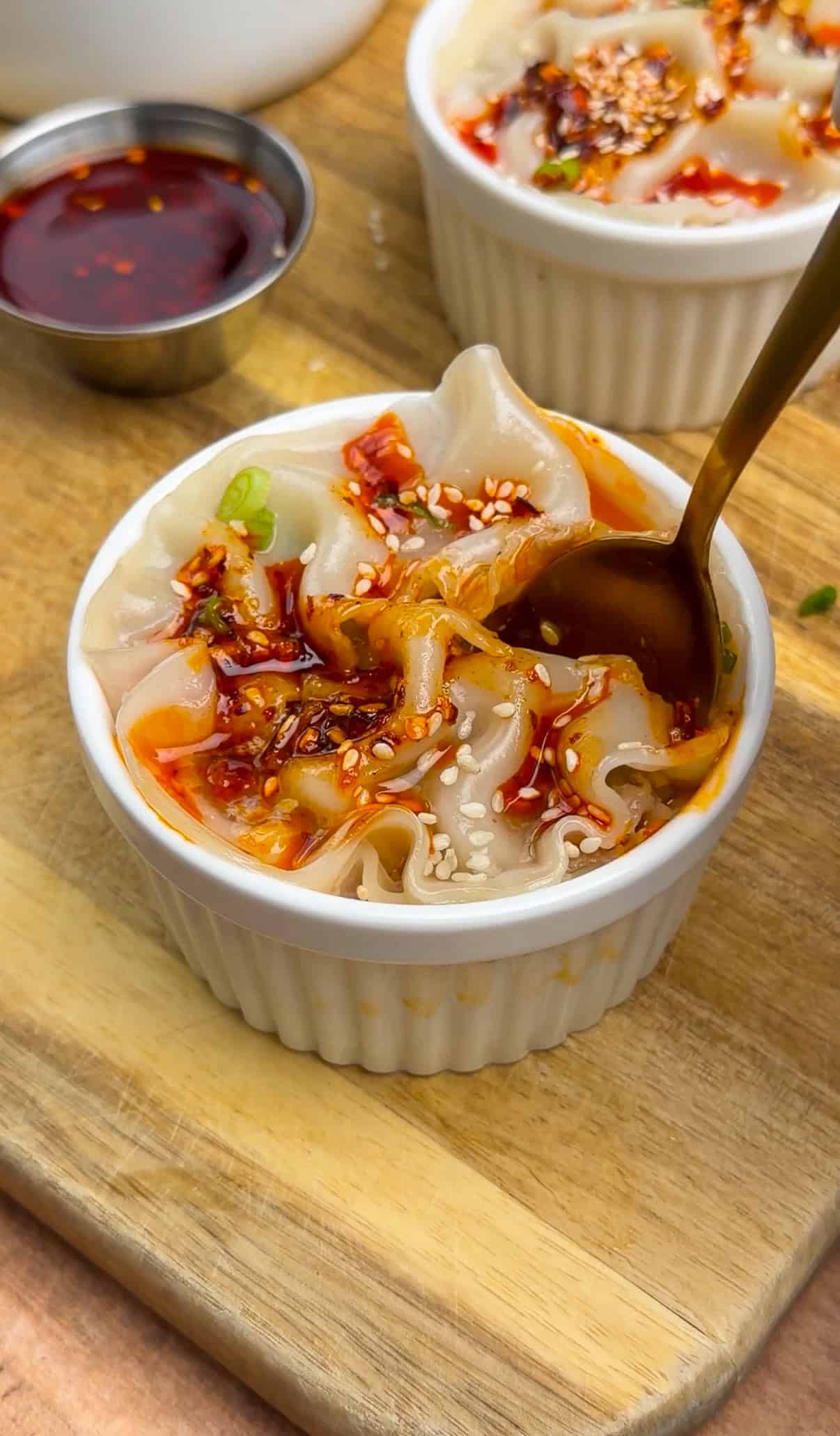 A white ramekin filled with dumplings topped with chili oil, sesame seeds, and green onions sits on a wooden board. A spoon is scooping a dumpling, and a small dish of chili oil is nearby. - 5
