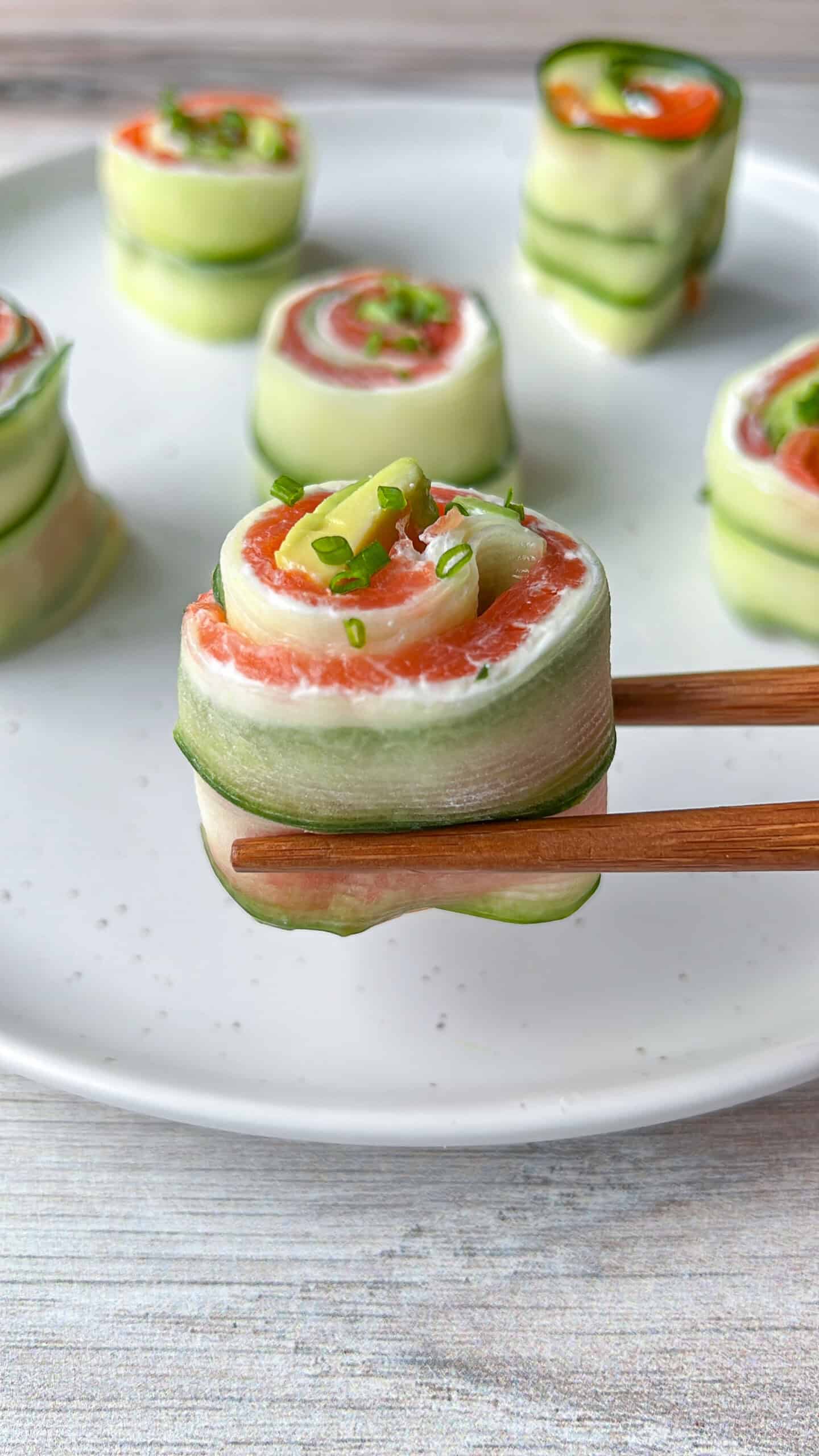 Cucumber-wrapped sushi rolls with salmon, cream cheese, and green onions are held by chopsticks above a white plate with more rolls in the background. - 3